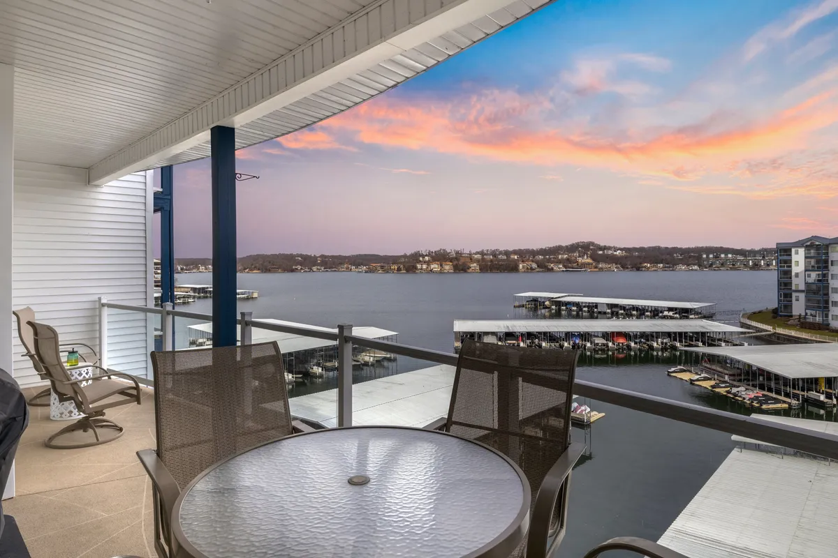 This exterior image presents a serene balcony space with outdoor furniture, a railing, and a sweeping lake view at sunset. The setup is ideal for al fresco dining, relaxation, and watching the sun dip below the horizon as a group enjoys the tranquil lakeside ambiance.