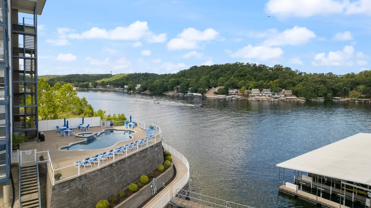 A vibrant outdoor scene featuring a sparkling swimming pool in the foreground, with the tranquil lake visible just beyond. Landscaped gardens surround the pool area, and several people are seen enjoying activities like swimming, sunbathing on lounge chairs, and relaxing by the water. A few boats are visible on the lake, emphasizing the cove-front location and recreational opportunities.