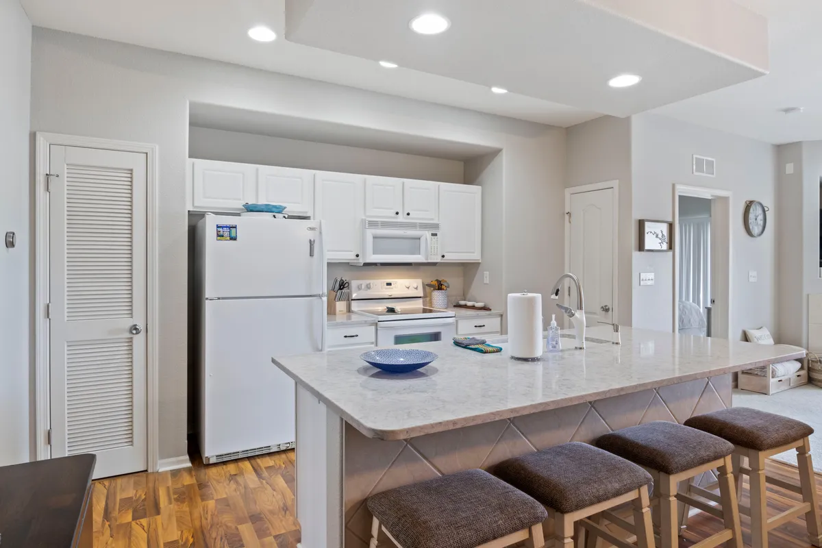 This image centers on a modern kitchen island with bar stools, surrounded by white cabinetry and countertop appliances. The arrangement is warm and welcoming—ideal for preparing meals or gathering for snacks and drinks. The visible details suggest a fully stocked, functional kitchen space ideal for families or groups.