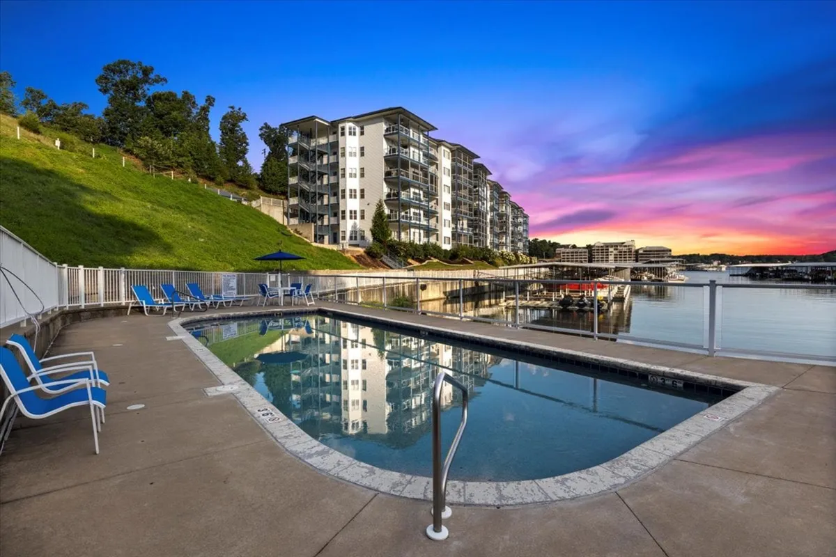 A sparkling swimming pool with reflections of the sunset in the water, surrounded by greenery and a modern apartment building. The sky glows with warm evening colors, emphasizing relaxation and picturesque views from the resort-style amenities.