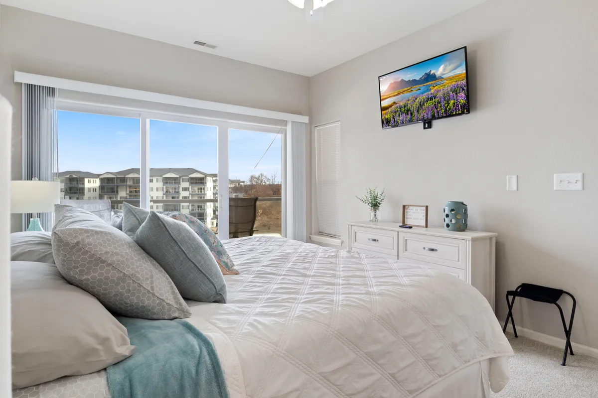 This image features a tranquil bedroom interior with a plush bed adorned with decorative pillows, a large window inviting in natural light, and a television mounted on the wall. The scene exudes coziness and serenity, suggesting a space perfect for unwinding, reading, or enjoying a quiet morning coffee.