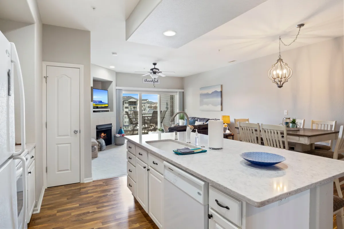 This image captures an open-concept interior with a kitchen island, dining area, and living room featuring a fireplace. Large windows reveal an outdoor space, allowing natural light and a view of the water to pour in. The space is arranged for both cooking and socializing, with inviting seating and a balance between practical amenities and homelike comfort.