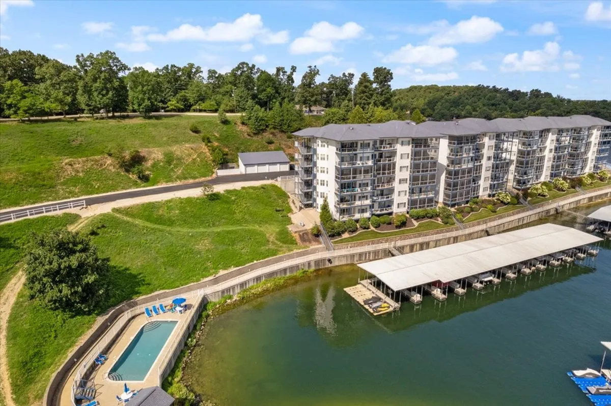 A serene lakeside setting with a prominent apartment building positioned beside a sparkling swimming pool and boat docks, lush greenery surrounding the water's edge. The wide dock area is clearly visible, offering direct access for boating while the pool and apartment evoke a resort-style ambiance.