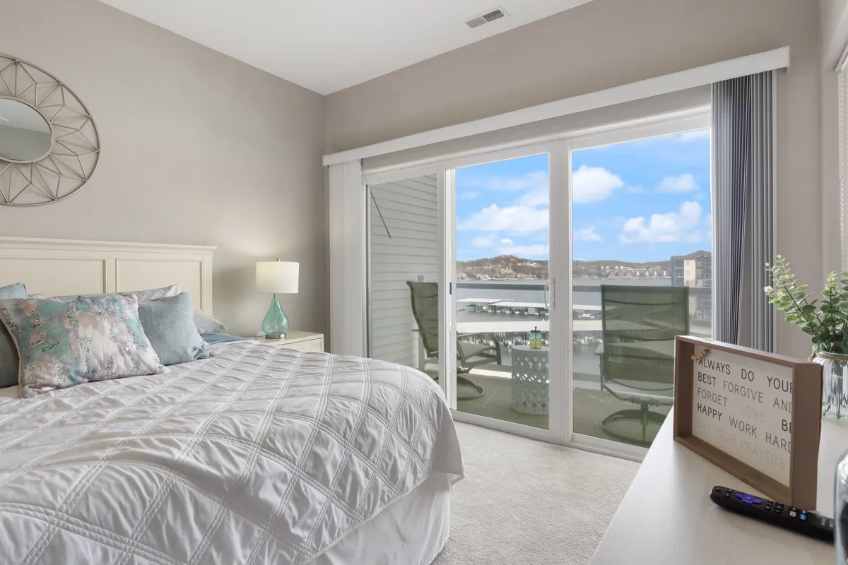 This serene bedroom scene includes a bed with decorative pillows, a sliding glass door leading to a balcony, a nightstand with a lamp and decor, and soft, natural light filling the space. The balcony suggests an extension of privacy and the opportunity to enjoy the outdoors in comfort.
