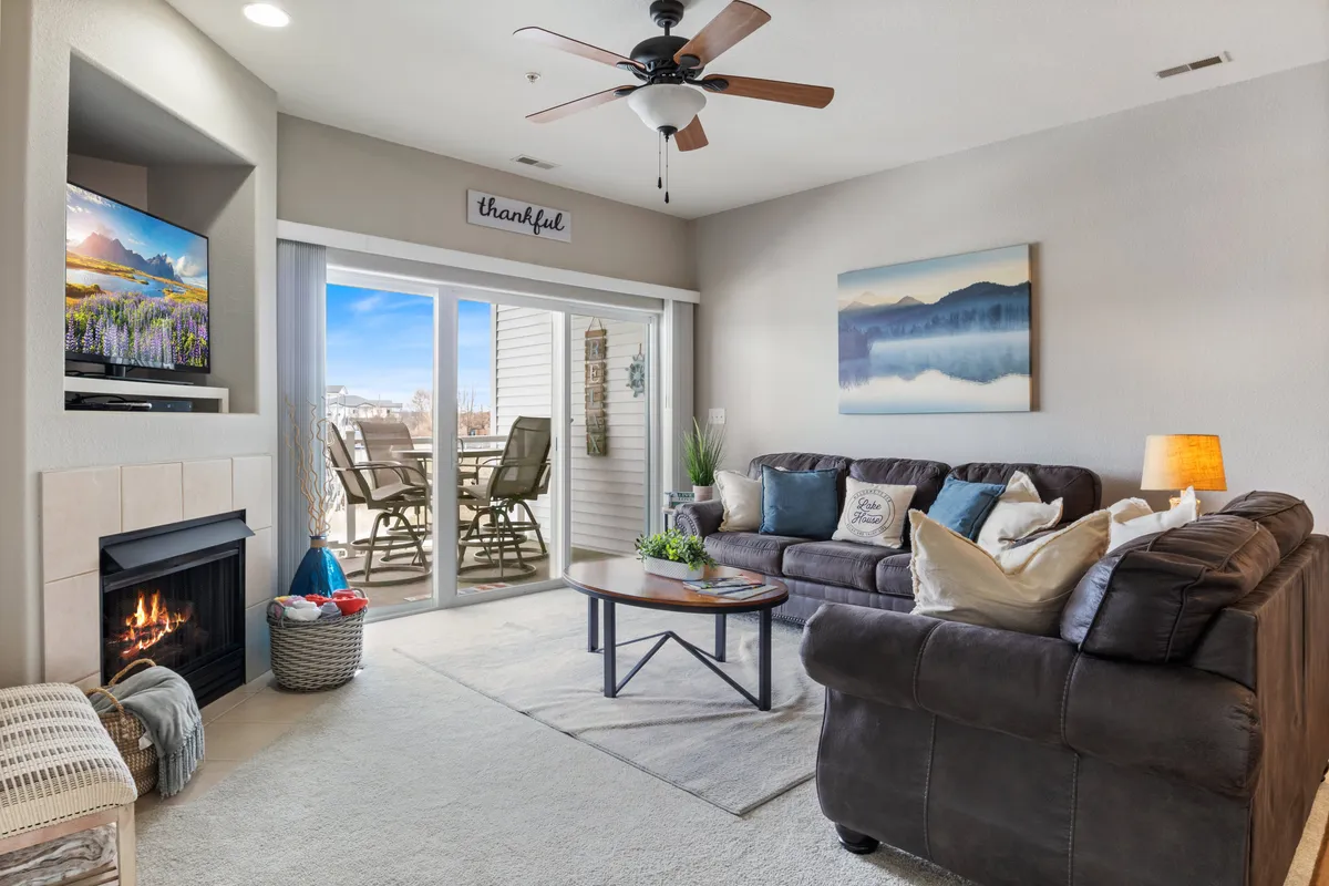 This interior image showcases a spacious, inviting living area featuring a stone fireplace, a large TV, a comfortable sofa set around a stylish coffee table, and a sliding glass door leading to a balcony with a stunning view of Lake Ozark. Natural light fills the room, and the balcony promises breathtaking lake views and serene mornings.