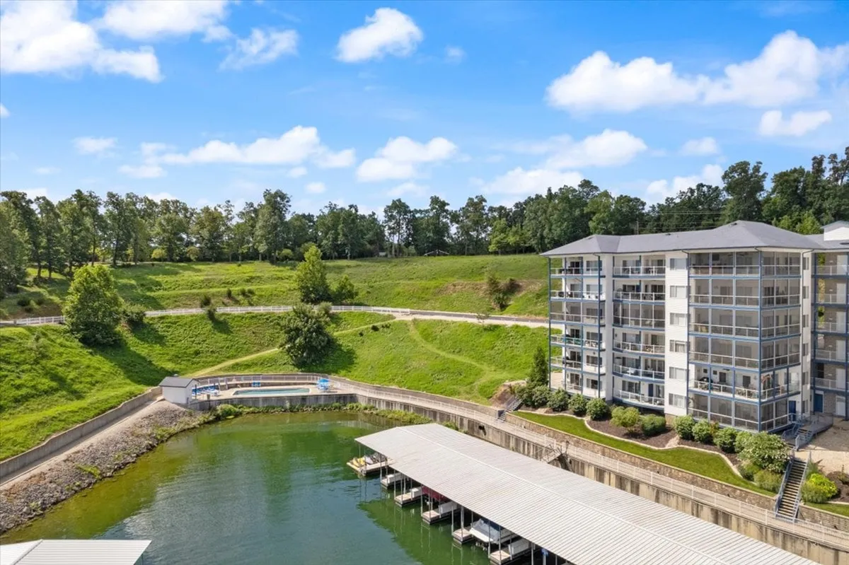 A tranquil lakeside setting with a wooden dock stretching out over a calm lake, framed by lush green landscaping and a modern apartment building in the background. The view is peaceful and inviting, perfect for quiet reflection, scenic strolls, or romantic moments by the water.