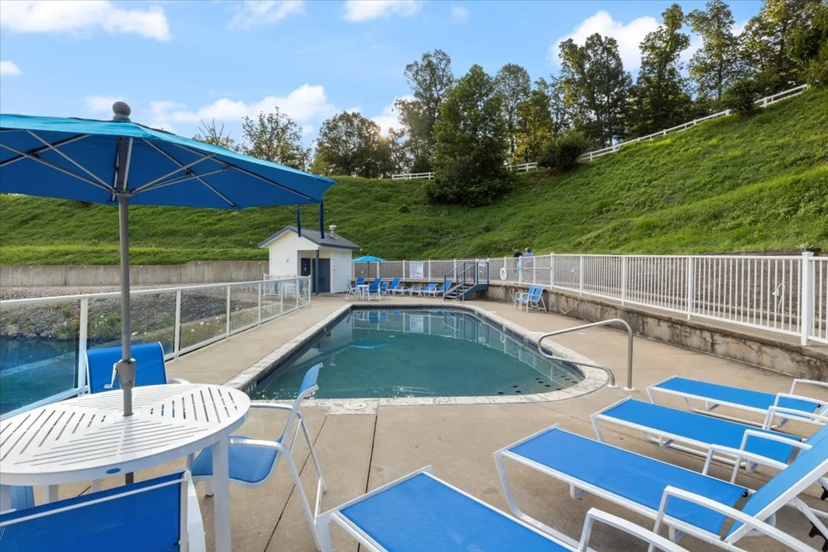 A sunny outdoor pool area with a clear blue swimming pool, lounge chairs, umbrellas, and a pool house. The inviting scene suggests a place for swimming, sunbathing, and family socializing, with a hint of the lake or waterfront nearby.